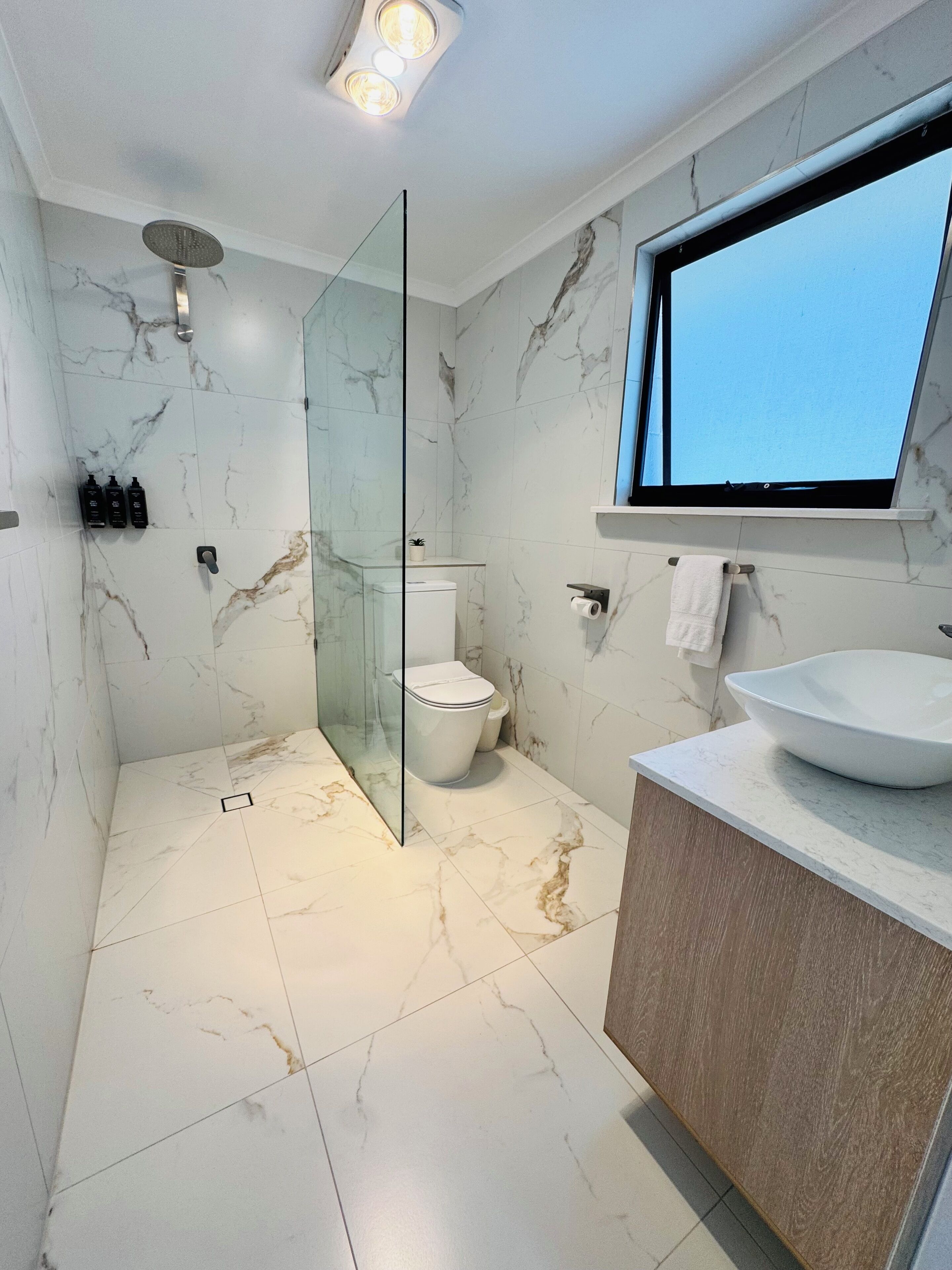 A modern bathroom with a glass-enclosed shower, a toilet, a sink with a large basin, and a window above the sink.
