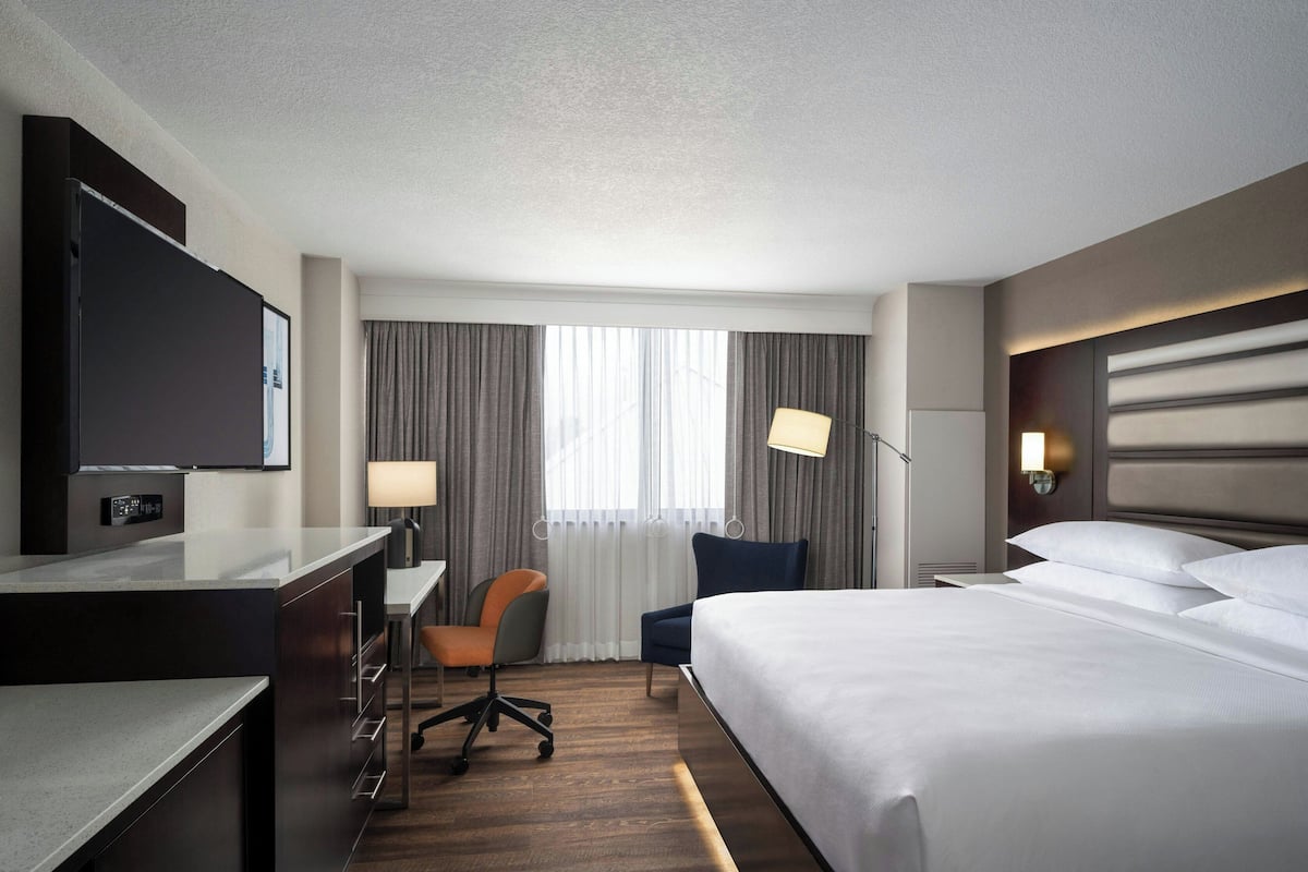 Una habitación de hotel con cama, escritorio, silla, televisor y ventana con cortinas.