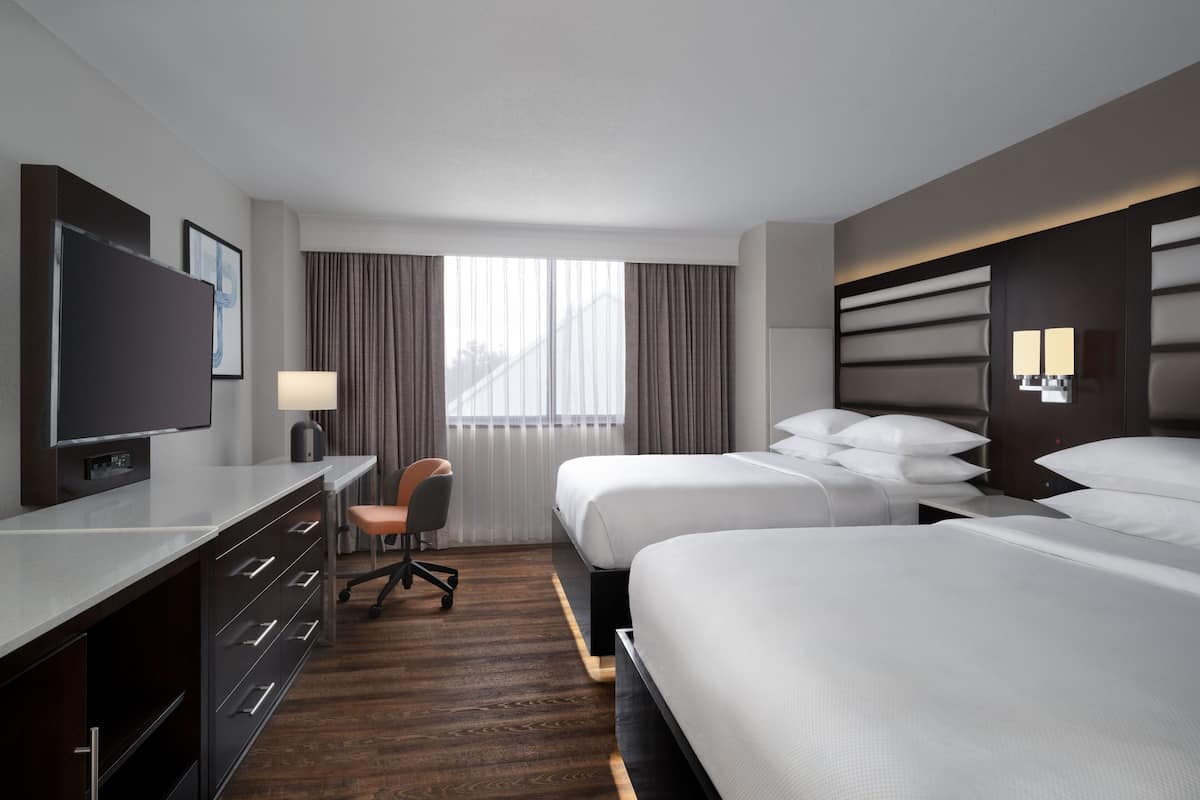 Una habitación de hotel con dos camas, un escritorio con silla, un televisor y un ventanal con cortinas.