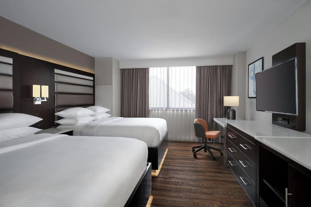 Habitación de hotel con dos camas, un escritorio con silla, televisor y ventana con cortinas.