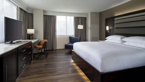Habitación de hotel con una cama grande, un escritorio, un televisor de pantalla plana, una silla y una ventana con cortinas.