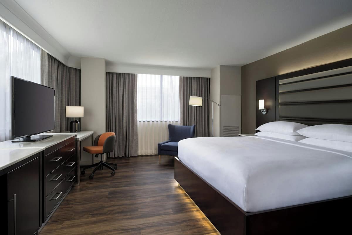 Habitación de hotel con una cama grande, un escritorio, un televisor de pantalla plana, una silla y una ventana con cortinas.