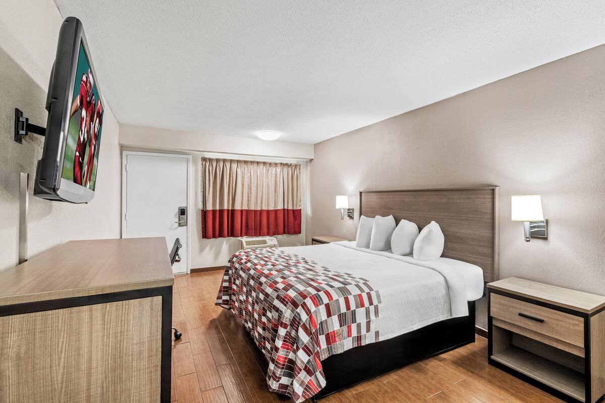 A hotel room with a bed, bedside tables, a desk, a TV, and a window with curtains.