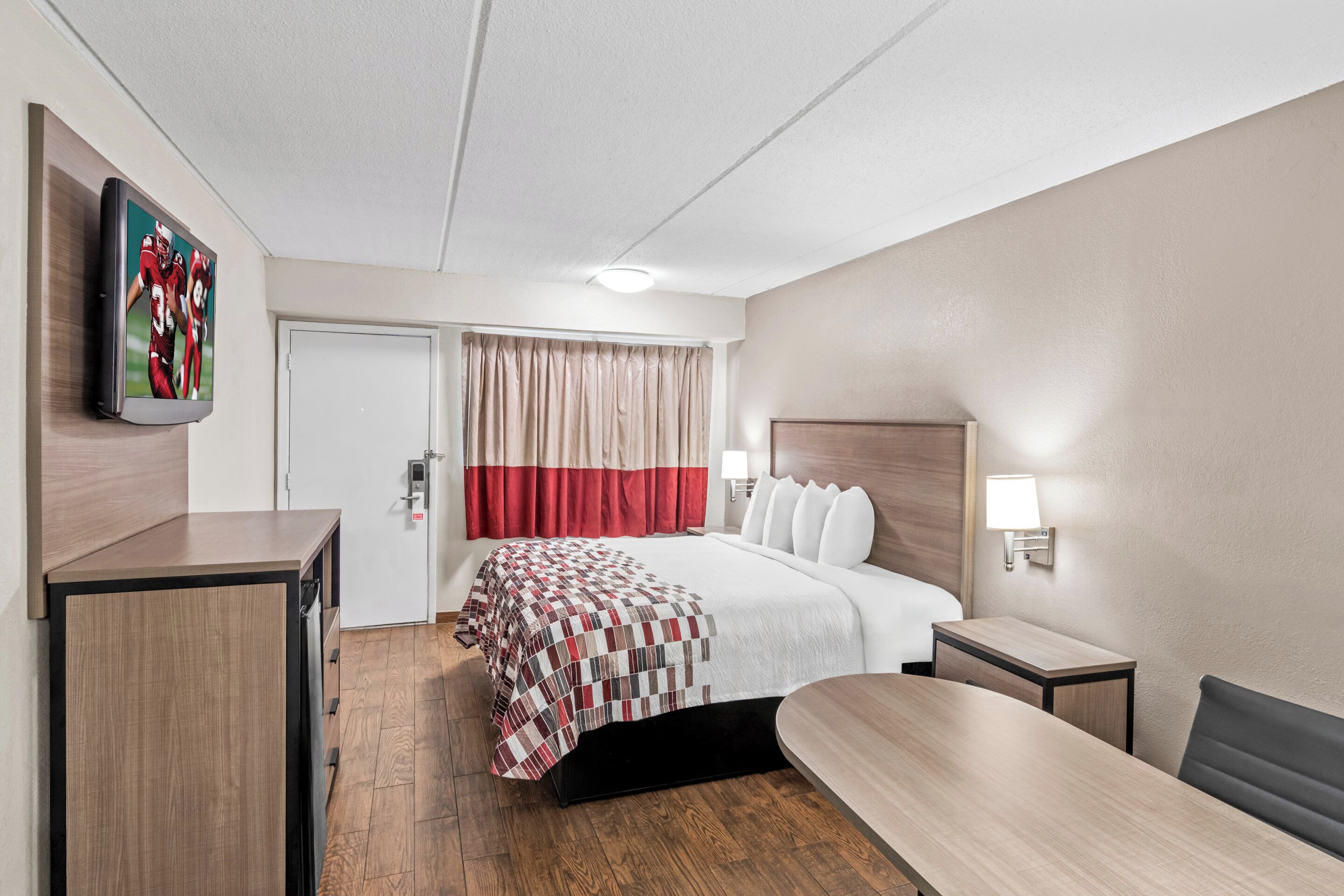 A hotel room with a bed, a desk, a chair, a TV mounted on the wall, and a window with curtains.