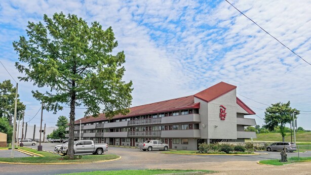 A multi-story building with a red roof, a parking lot, and a large tree in front. - Red Roof Inn Jackson Downtown - Fairgrounds (Jackson)