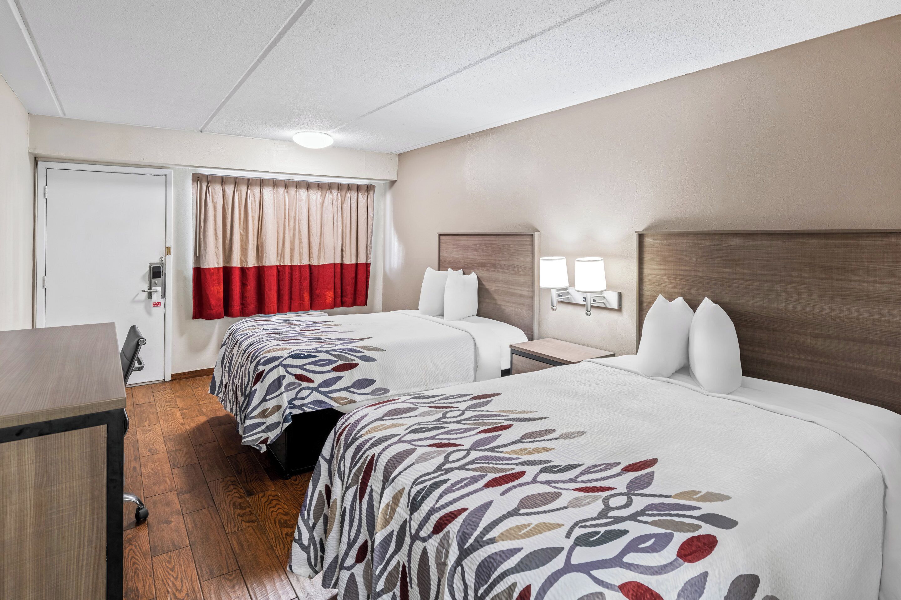 Habitación de hotel con dos camas, un escritorio, una silla, una puerta y una ventana con cortinas.