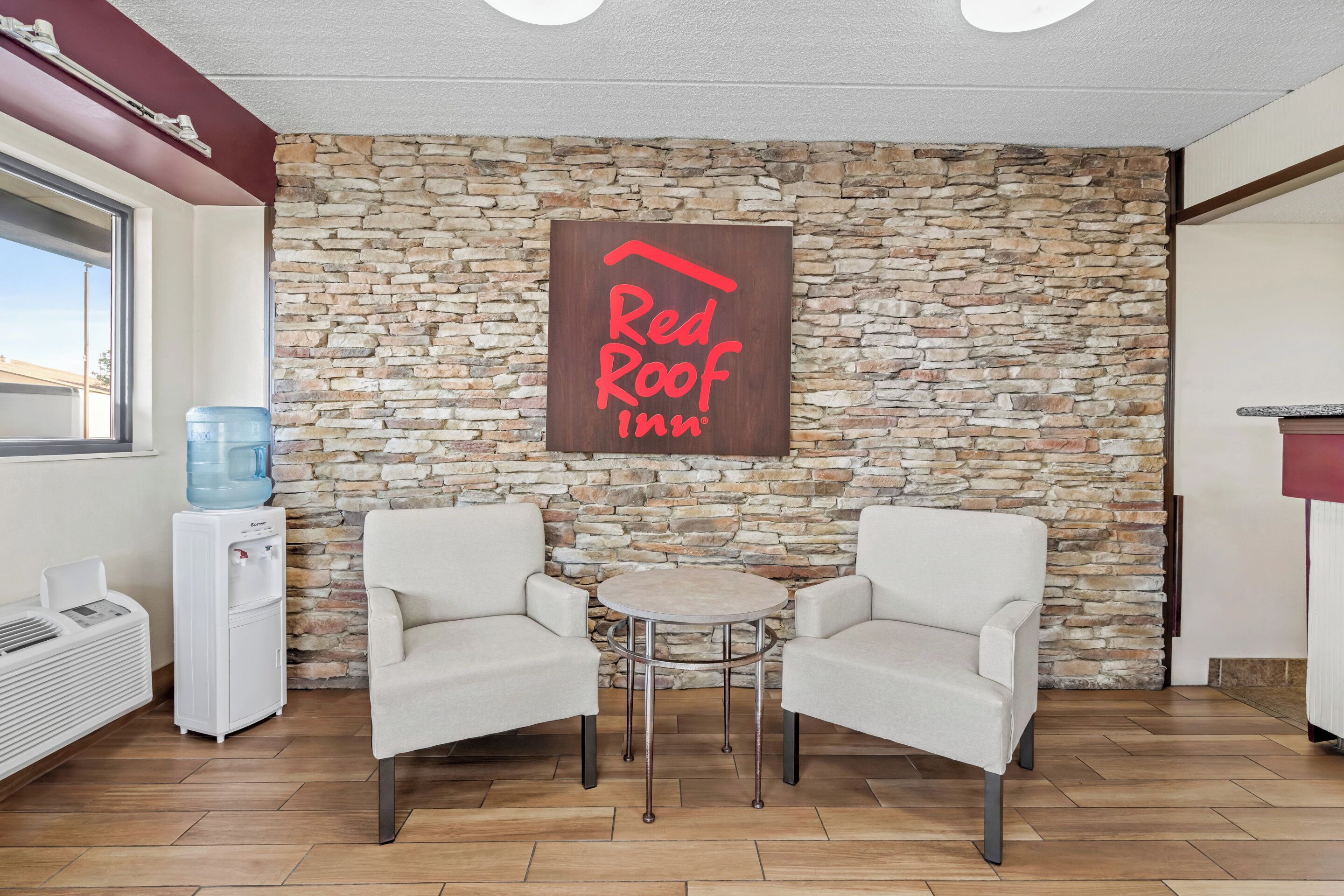 Quarto Red Roof Inn com parede de pedra, dois poltronas brancas, uma mesa redonda pequena, um bebedouro e uma janela.