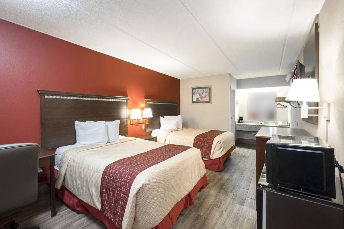 A hotel room with two beds, a desk, a television, and a bathroom visible in the background.