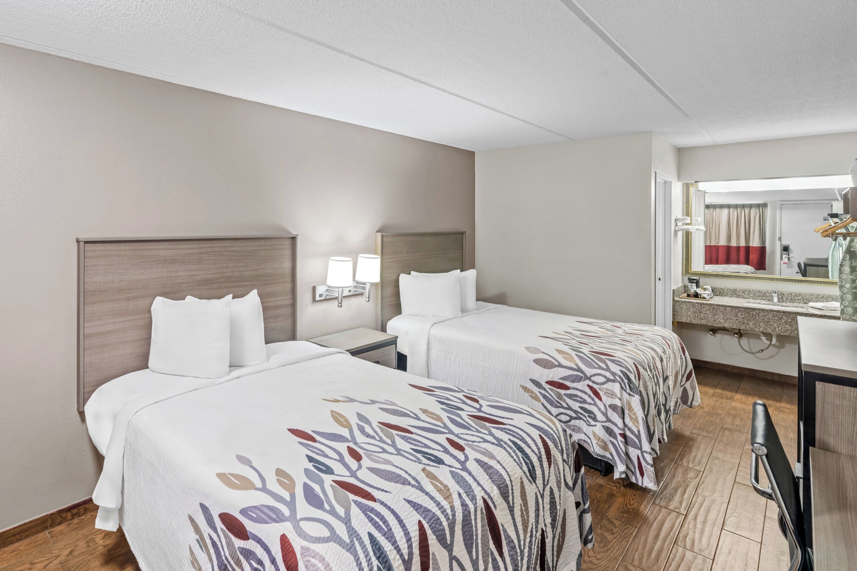 Une chambre d’hôtel avec deux lits, une table de chevet avec des lampes, un miroir et une salle de bain visible à travers une porte ouverte.