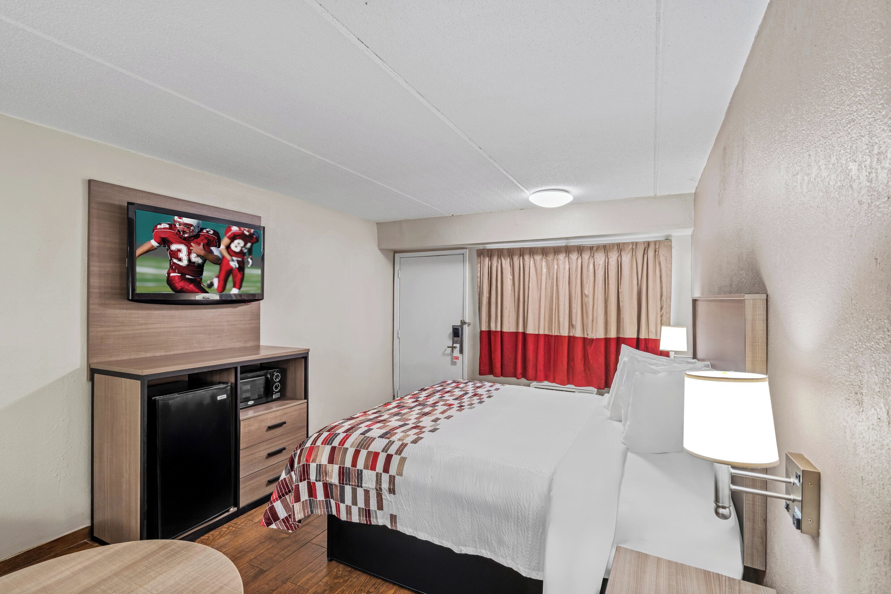 A hotel room with a bed, a TV mounted on the wall, a desk with a microwave, and a window with curtains.
