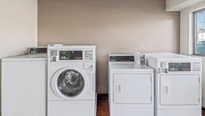 A laundry area with three stacked washers and dryers, beige walls, and a window. - Red Roof Inn Jackson Downtown - Fairgrounds (Jackson)