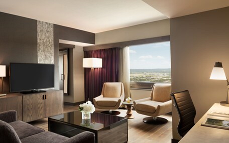 A modern hotel room with a flat-screen TV, two armchairs, a sofa, a coffee table, and a large window offering a city view.