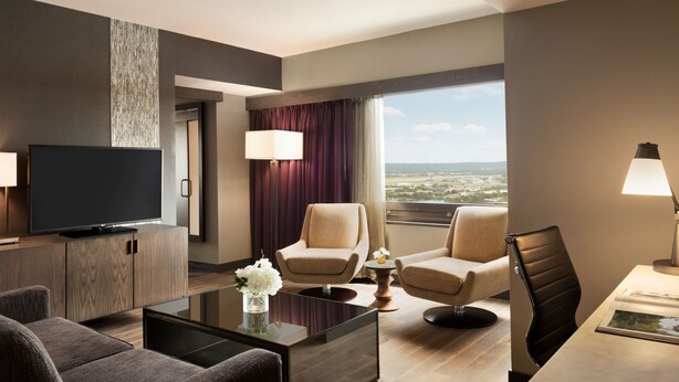 Una habitación de hotel moderna con televisión de pantalla plana, dos sillones, un sofá, una mesita de centro y un ventanal con vistas a la ciudad.