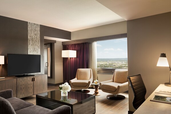 Een moderne hotelkamer met een flatscreen televisie, twee fauteuils, een bank, een salontafel en een groot raam met uitzicht op de stad.
