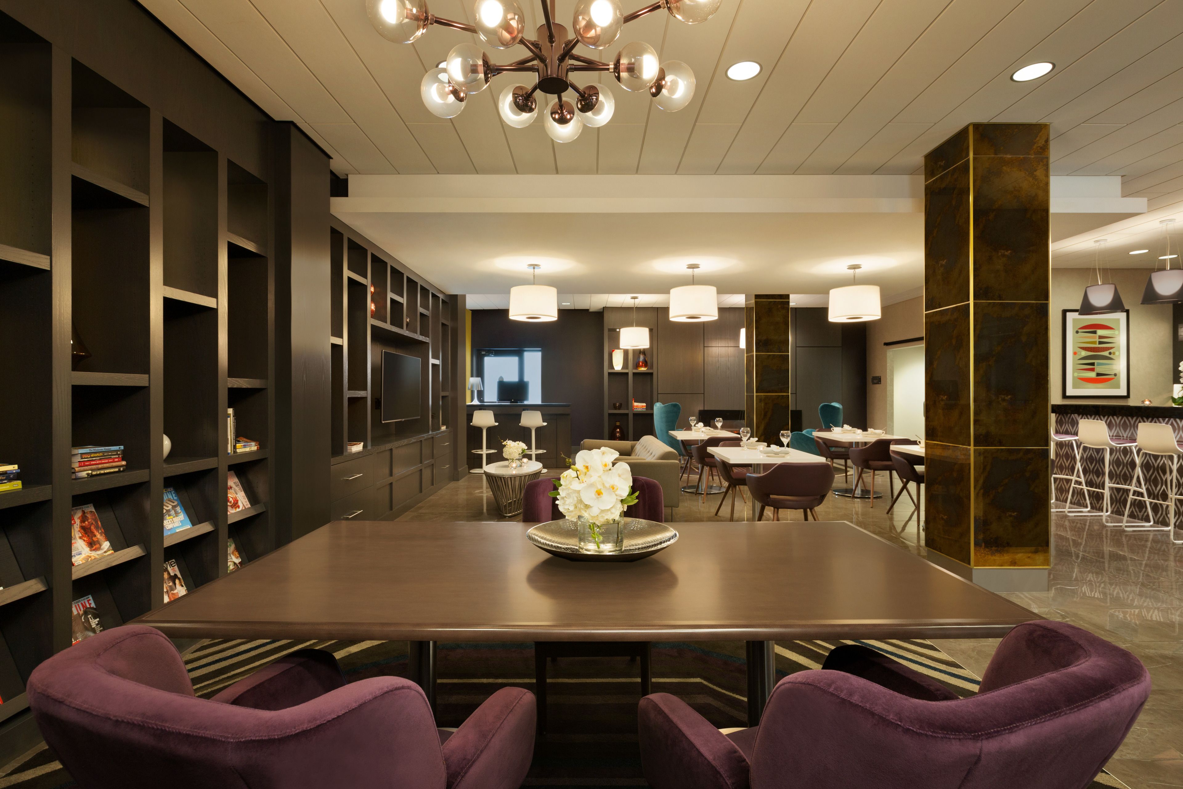A modern dining area with a long table, purple chairs, and a chandelier. There are bookshelves and a bar area in the background.