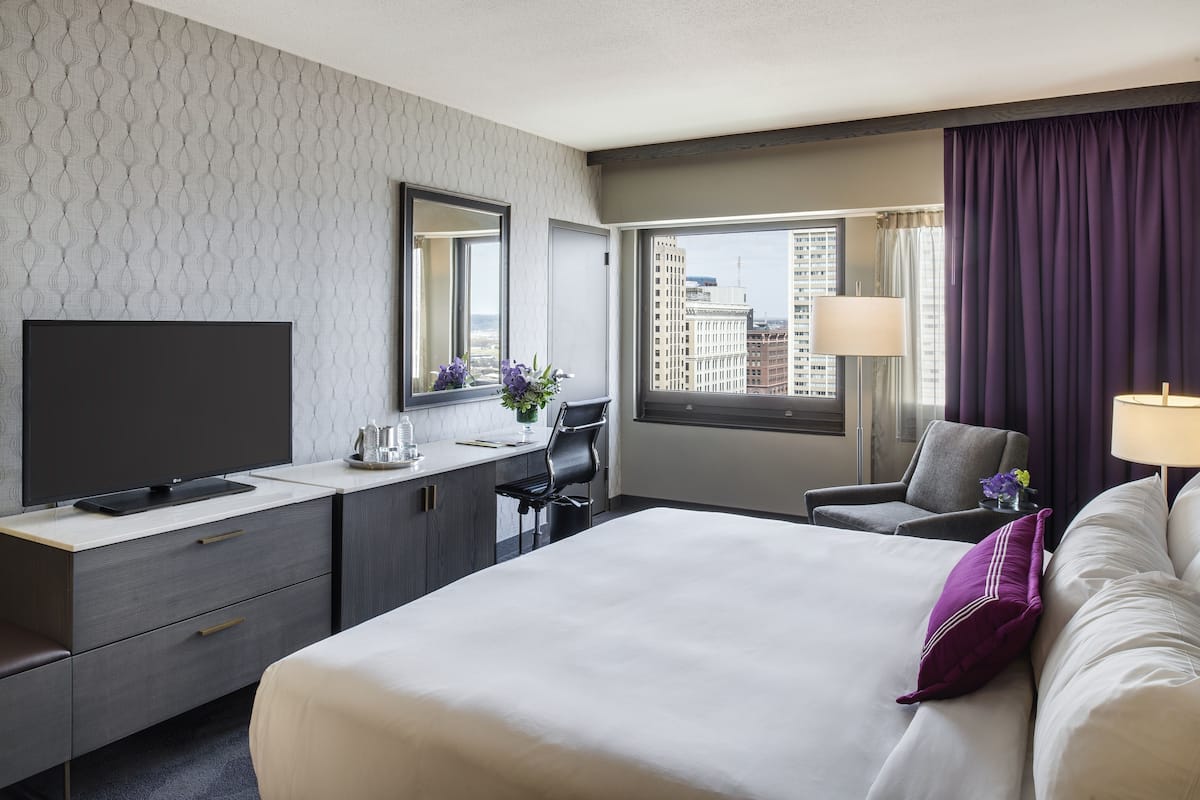 A modern hotel room with a large bed, a desk with a flat-screen TV, a chair, and a window with a city view.