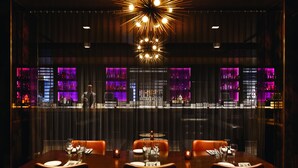 A dimly lit bar area with a modern chandelier, a bar counter with purple lighting, and a dining area with tables set for two.