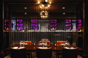 Zona de bar con iluminación tenue, un candelabro moderno, una barra con luces moradas y un área de comedor con mesas para dos.