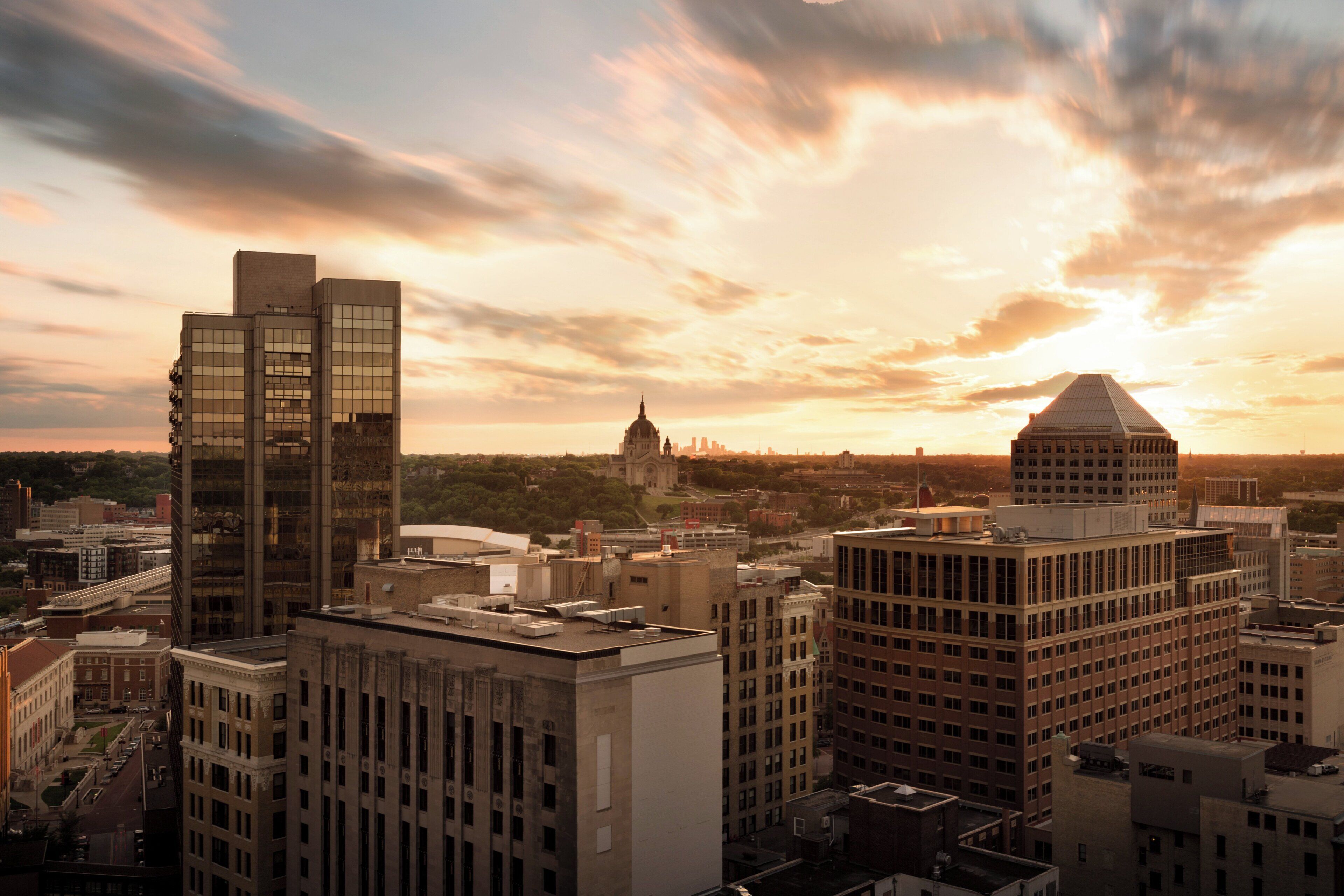 A cityscape with modern buildings, including a prominent skyscraper and a historic church, under a sunset sky.