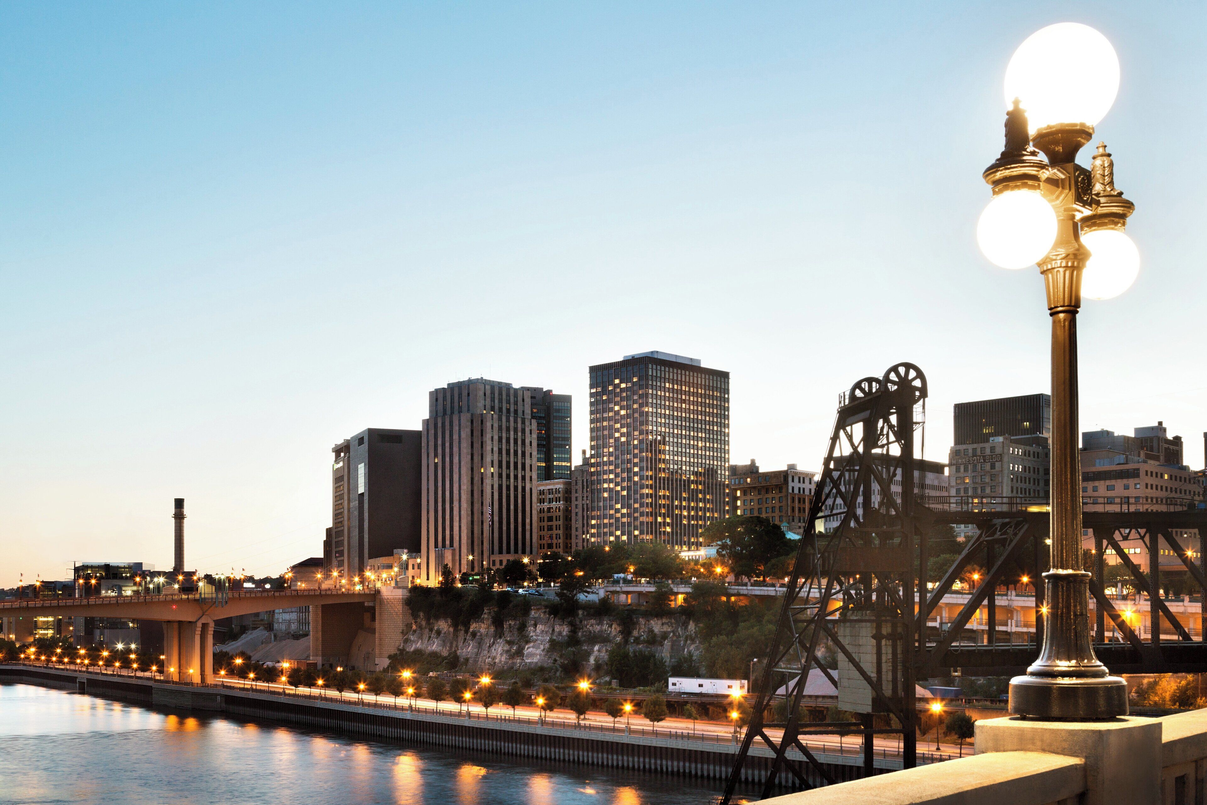 A city skyline with modern buildings, a river, and a bridge with a street lamp.