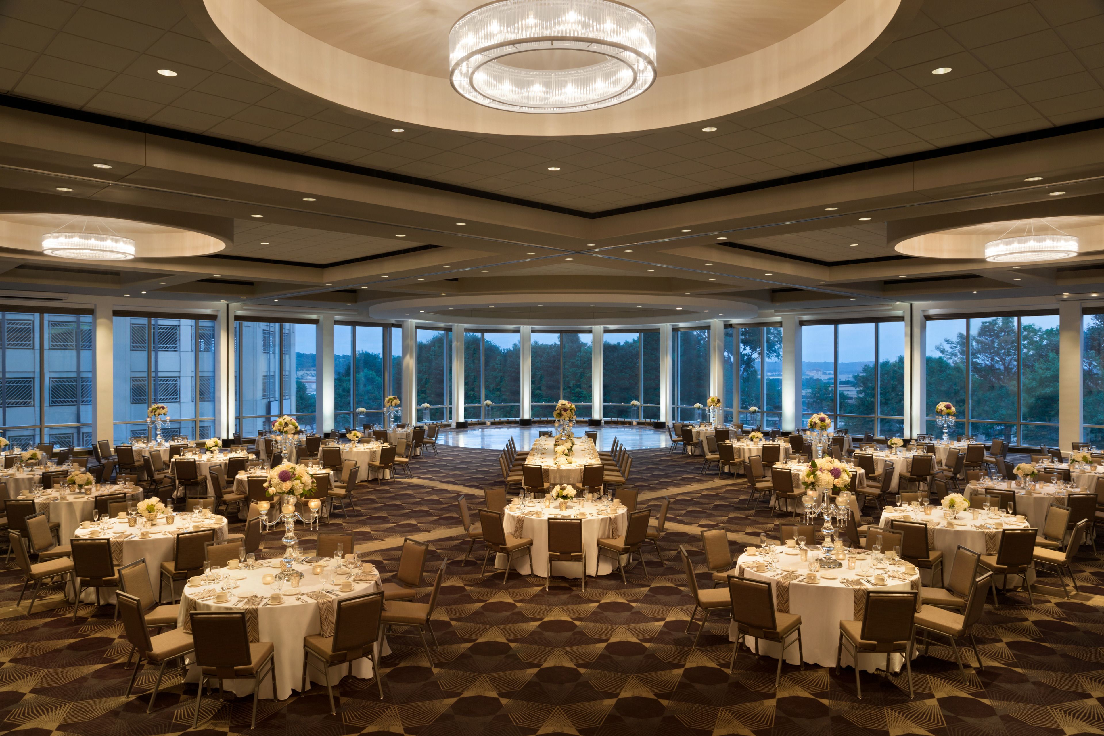 A spacious banquet hall with round tables, chandeliers, and large windows offering a view of the outdoors.