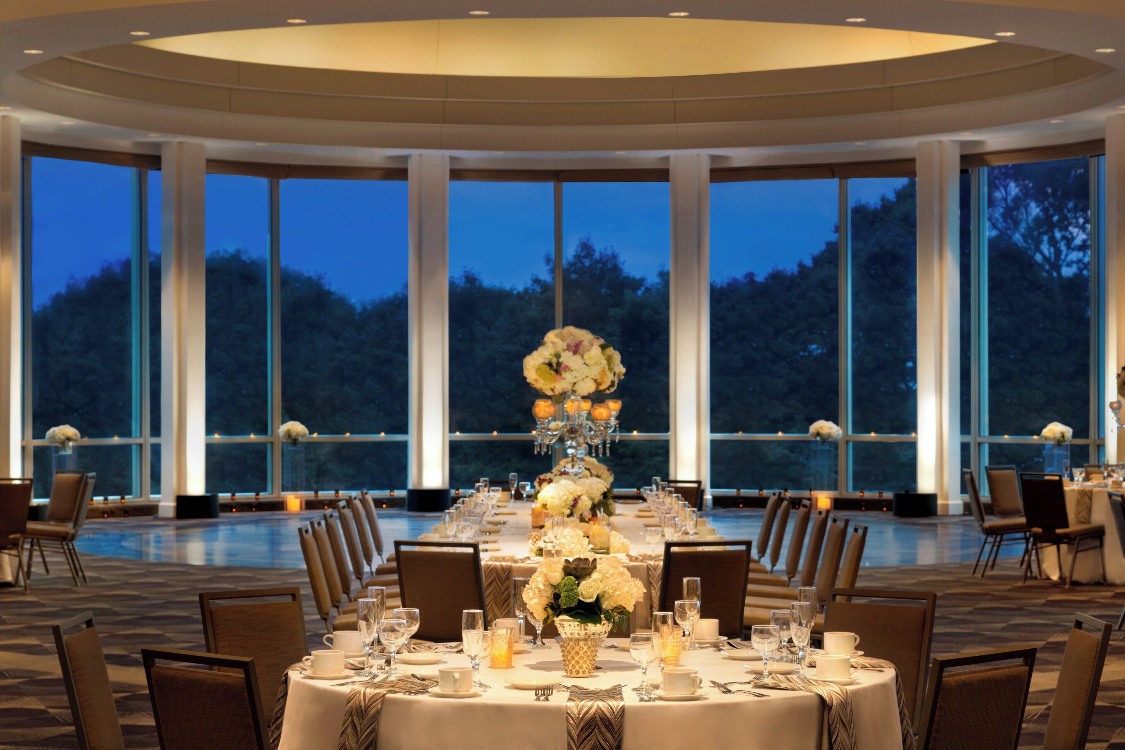 Un comedor formal con una mesa grande dispuesta para una comida, rodeada de sillas y grandes ventanales que ofrecen vistas al exterior.