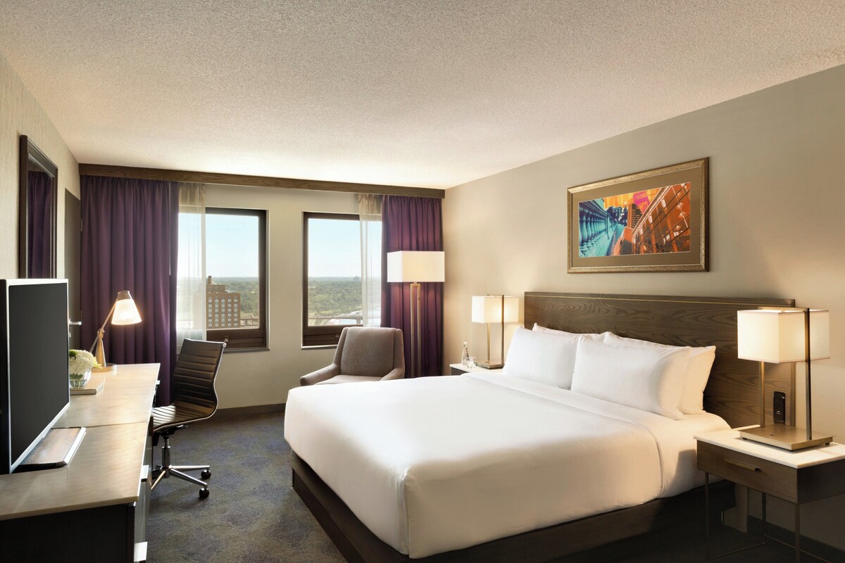 A hotel room with a large bed, a desk with a computer, a chair, two windows with curtains, and a painting on the wall.