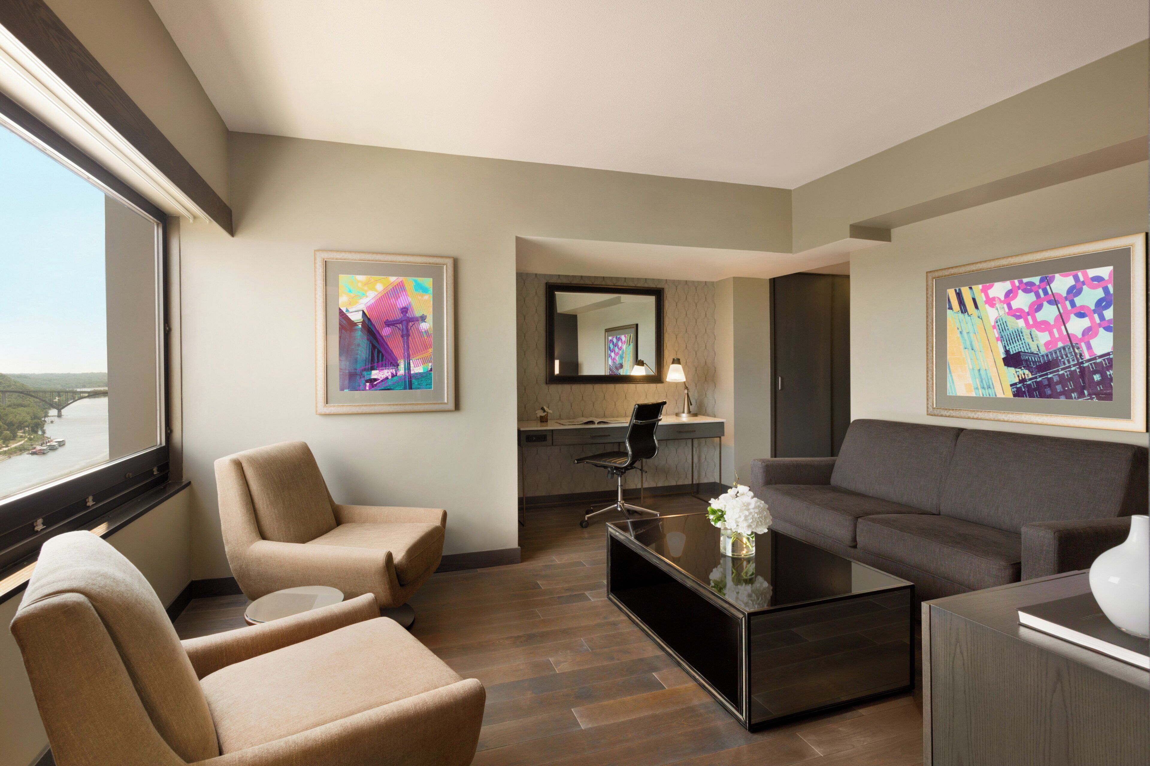 A hotel room with a sofa, armchairs, a coffee table, a desk, and a large window offering a view of a bridge.