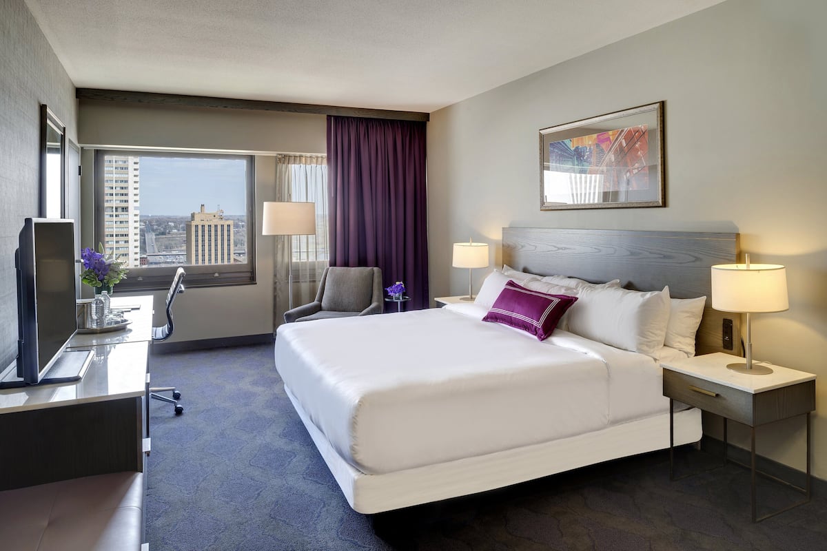 A hotel room with a large bed, a desk, a chair, a TV, and a window with a city view.