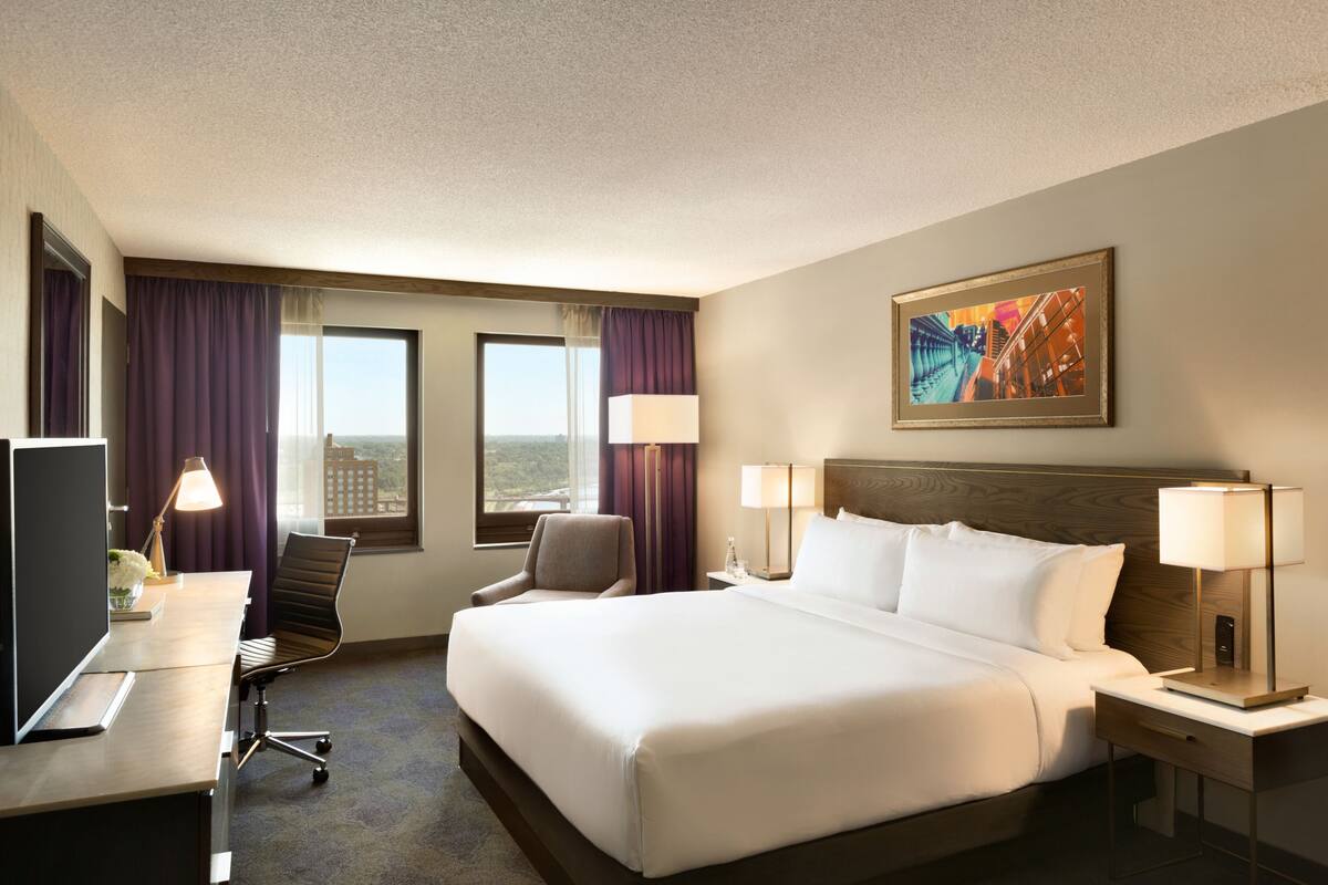 A hotel room with a large bed, a desk with a computer, a chair, two windows with curtains, and a painting on the wall.