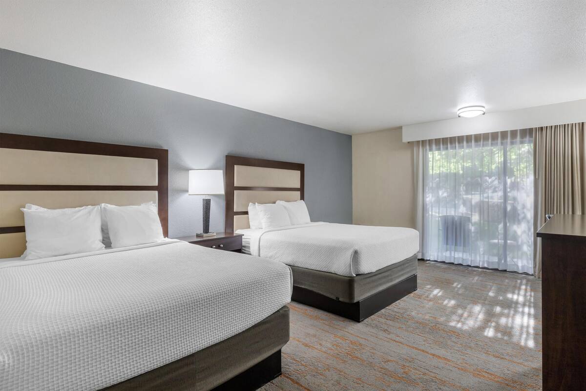 A hotel room with two beds, a desk, a lamp, and a large window with curtains.