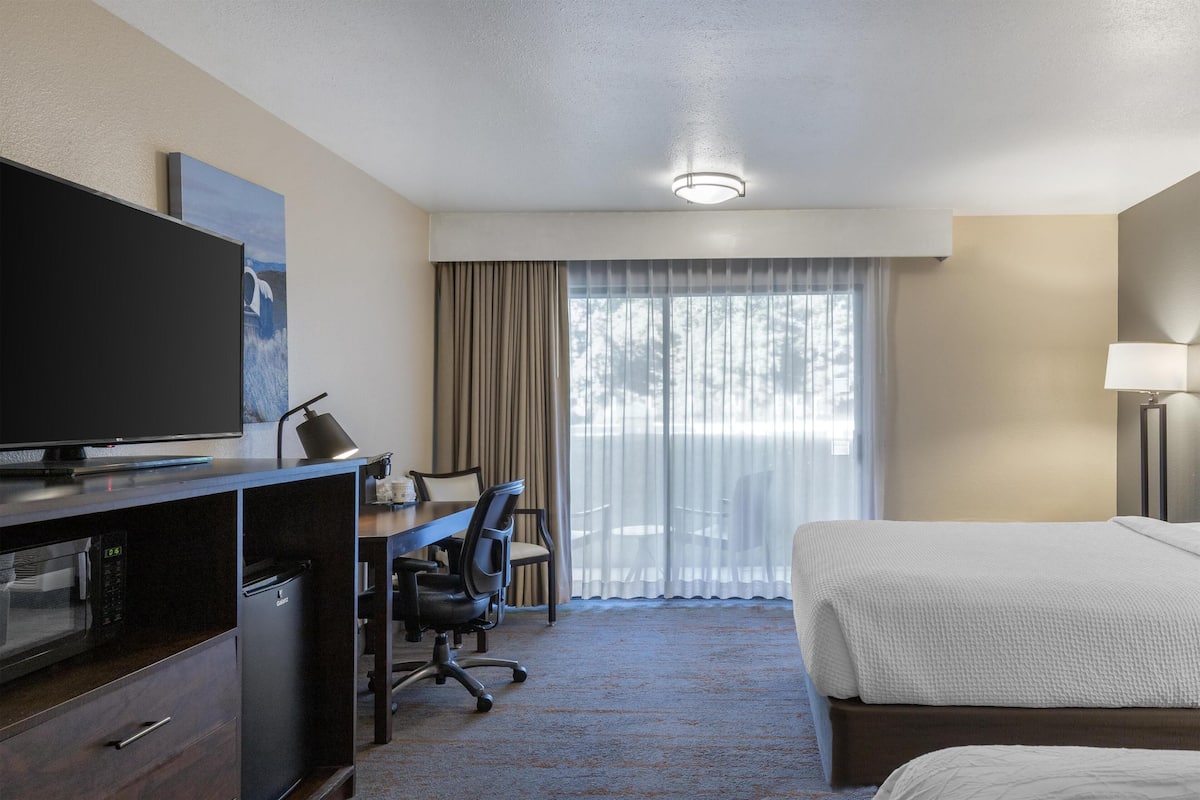 A hotel room with a bed, a desk with a chair, a television, a microwave, and a large window with curtains.