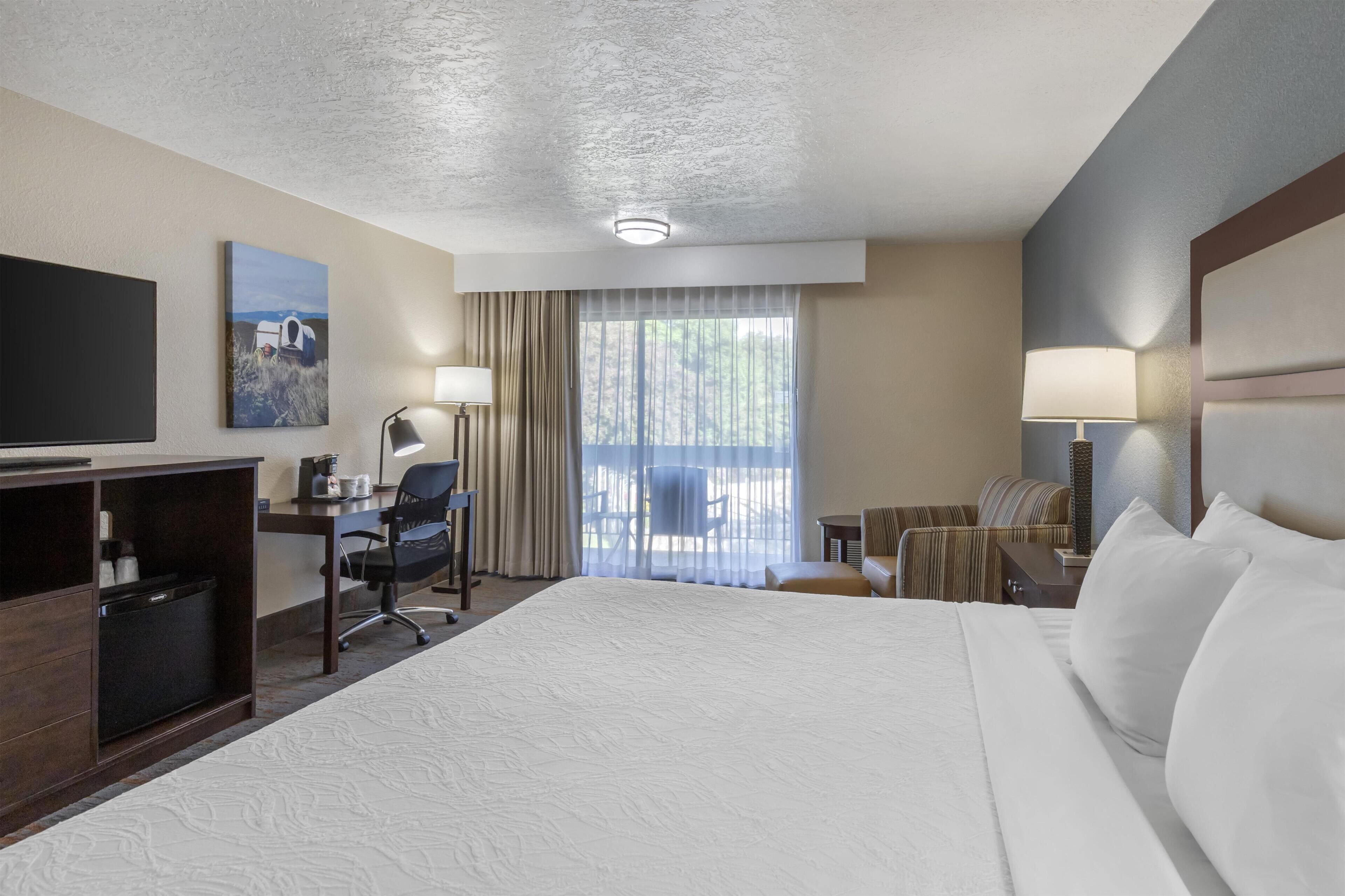 Quarto de hotel com uma cama grande, uma escrivaninha com cadeira, televisão e janela com cortinas.