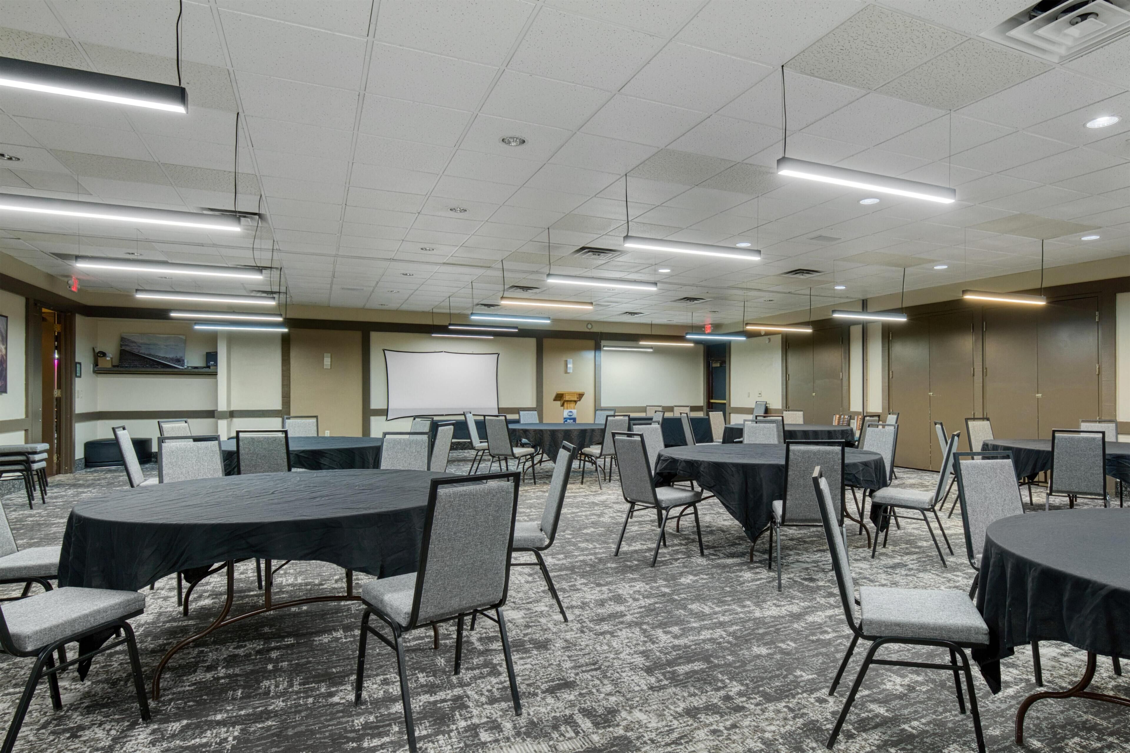 Une salle de conférence dotée de plusieurs tables et chaises, d’un écran de projection et d’un sol recouvert de moquette.