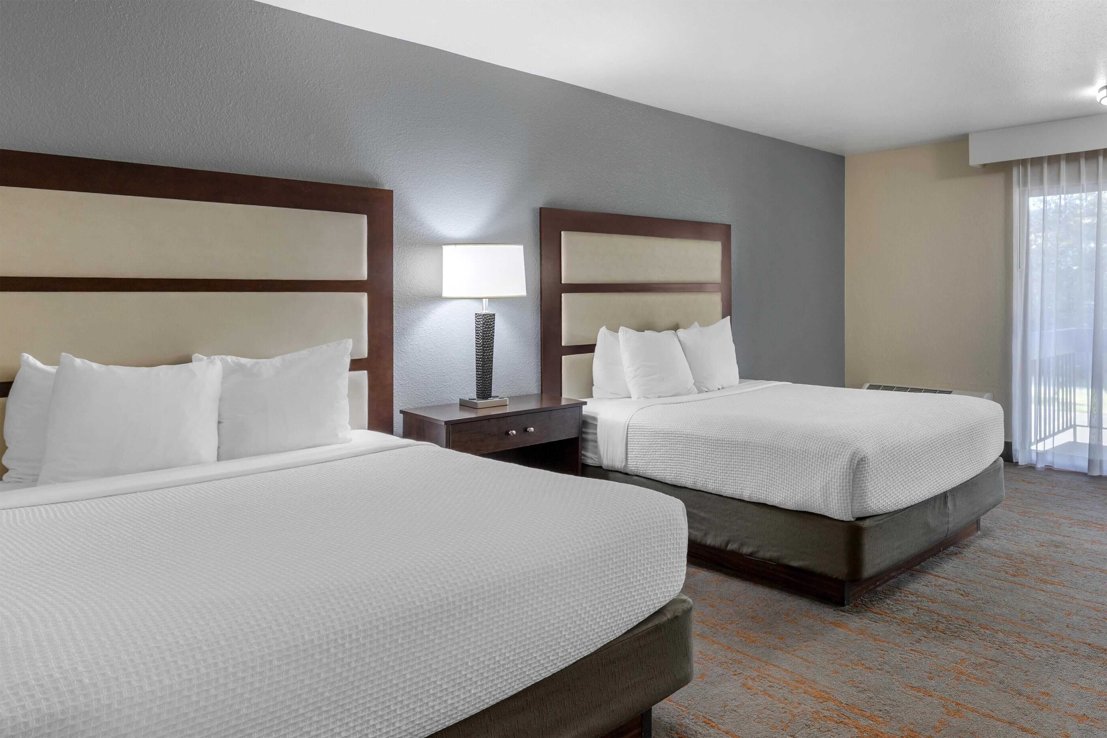 A hotel room with two beds, a nightstand, a lamp, and a large window with curtains.