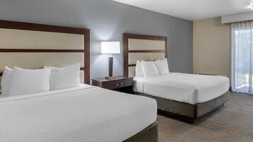 A hotel room with two beds, a nightstand, a lamp, and a large window with curtains.