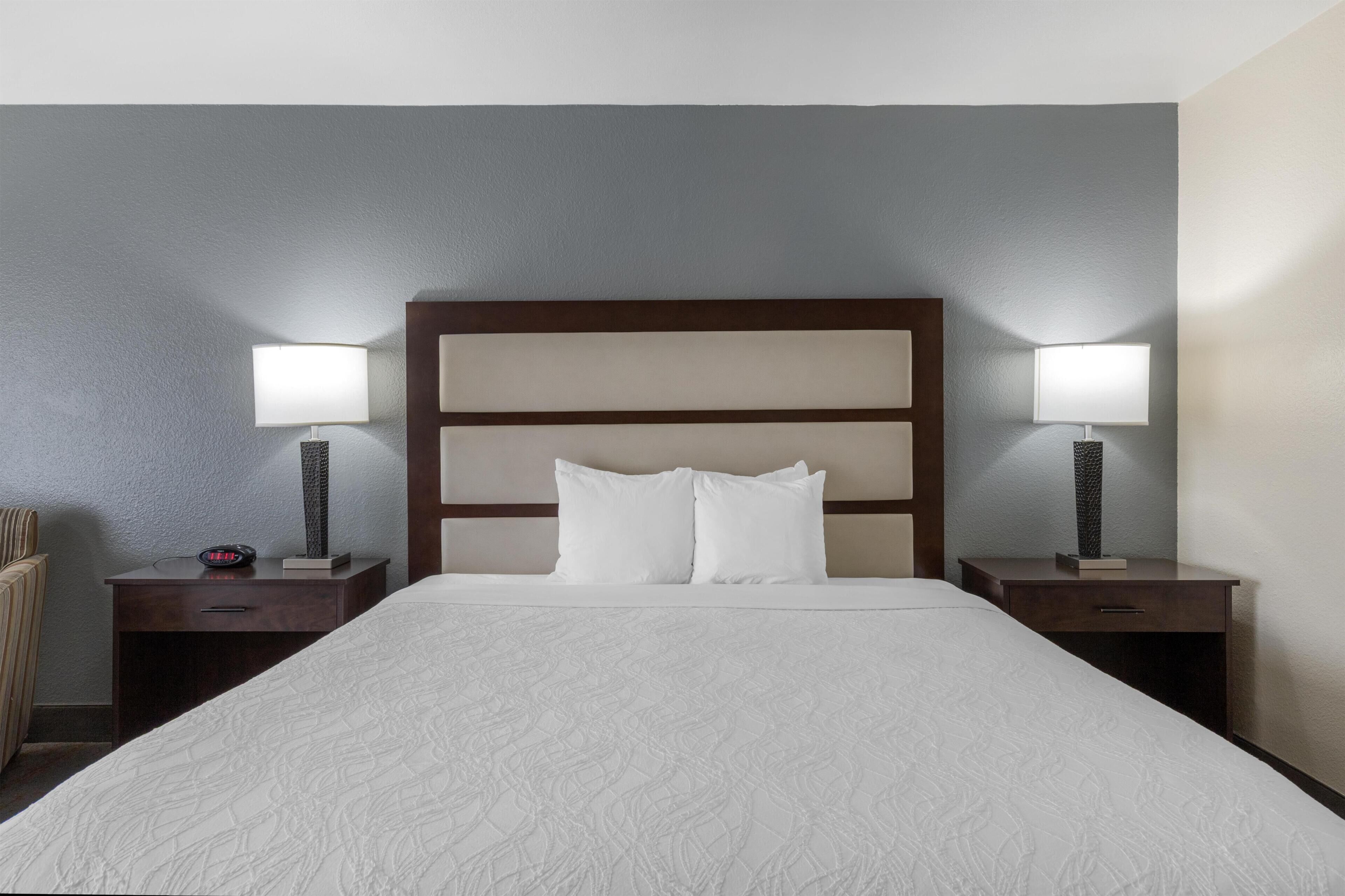A hotel room with a large bed, two bedside tables with lamps, a clock, and a chair.