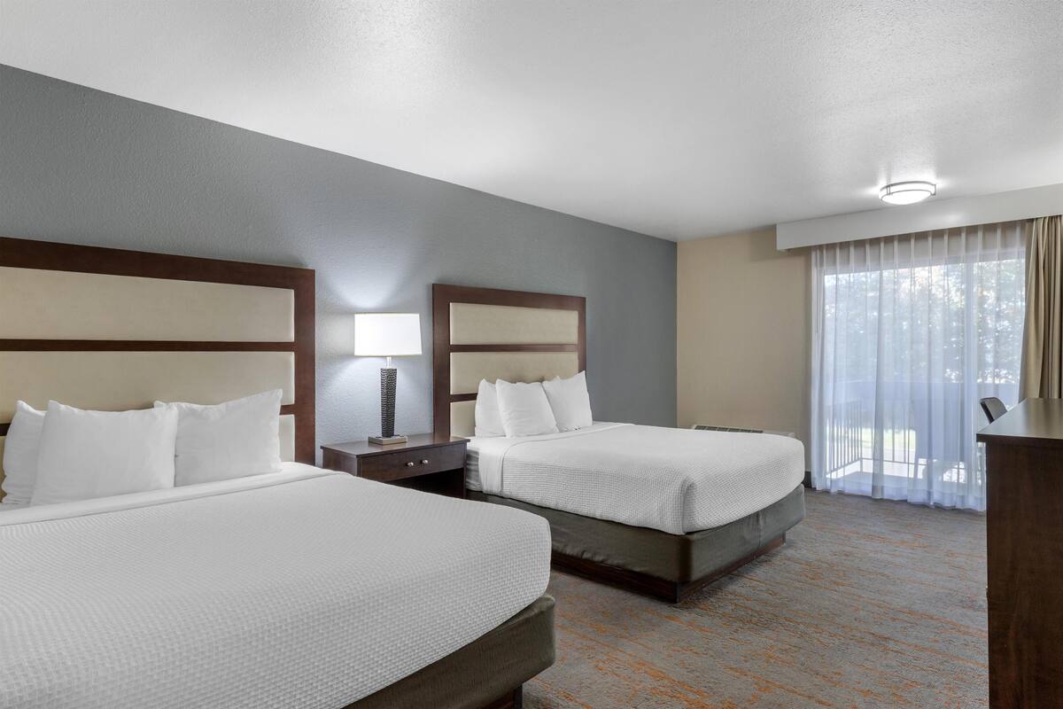 A hotel room with two beds, a nightstand, a lamp, and a view of the outdoors through a window.