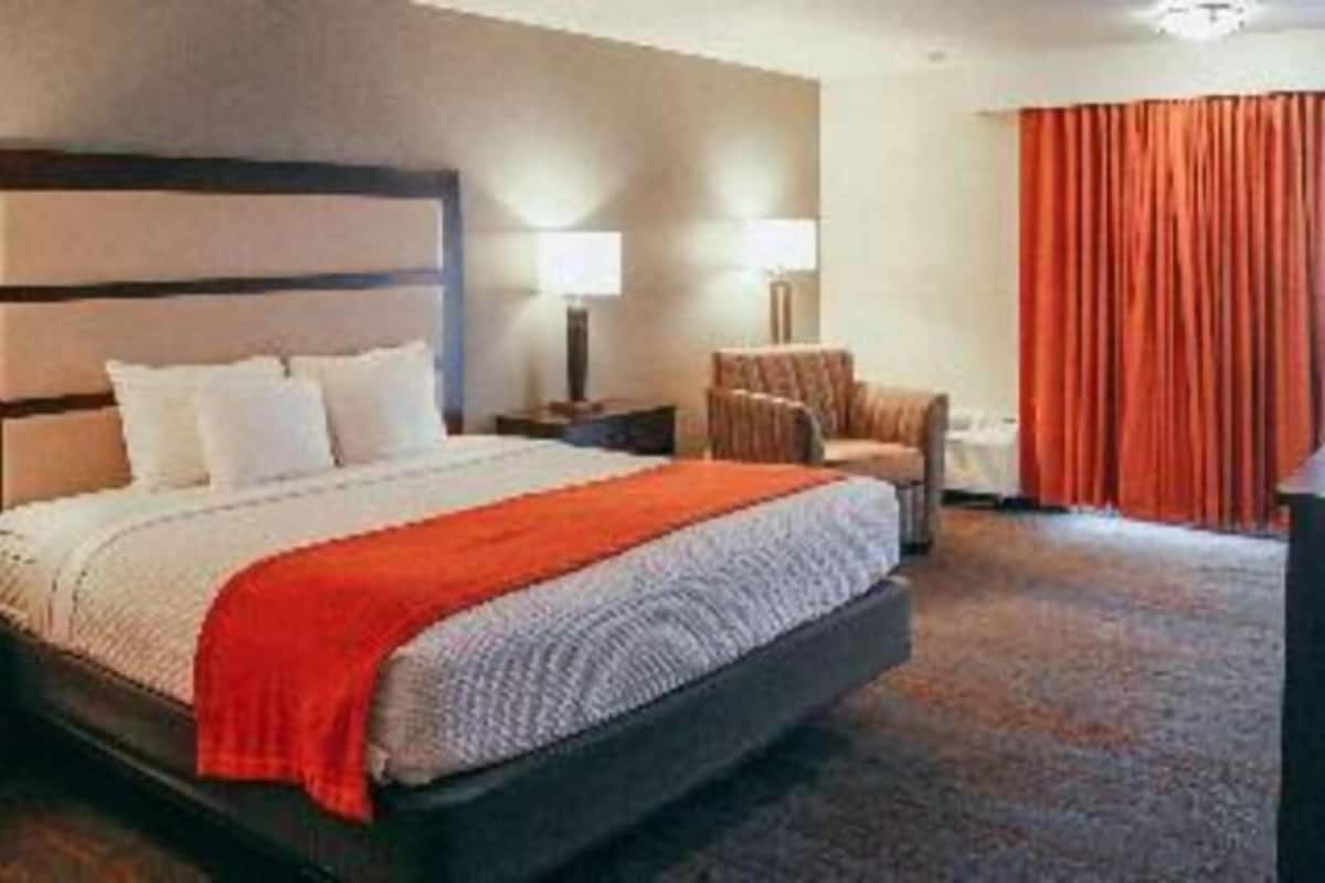 Une chambre d’hôtel avec un lit, une chaise, une table de chevet, une lampe et une fenêtre avec des rideaux.