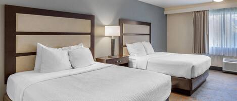 A hotel room with two beds, a nightstand with a lamp, a window with curtains, and a ceiling light.