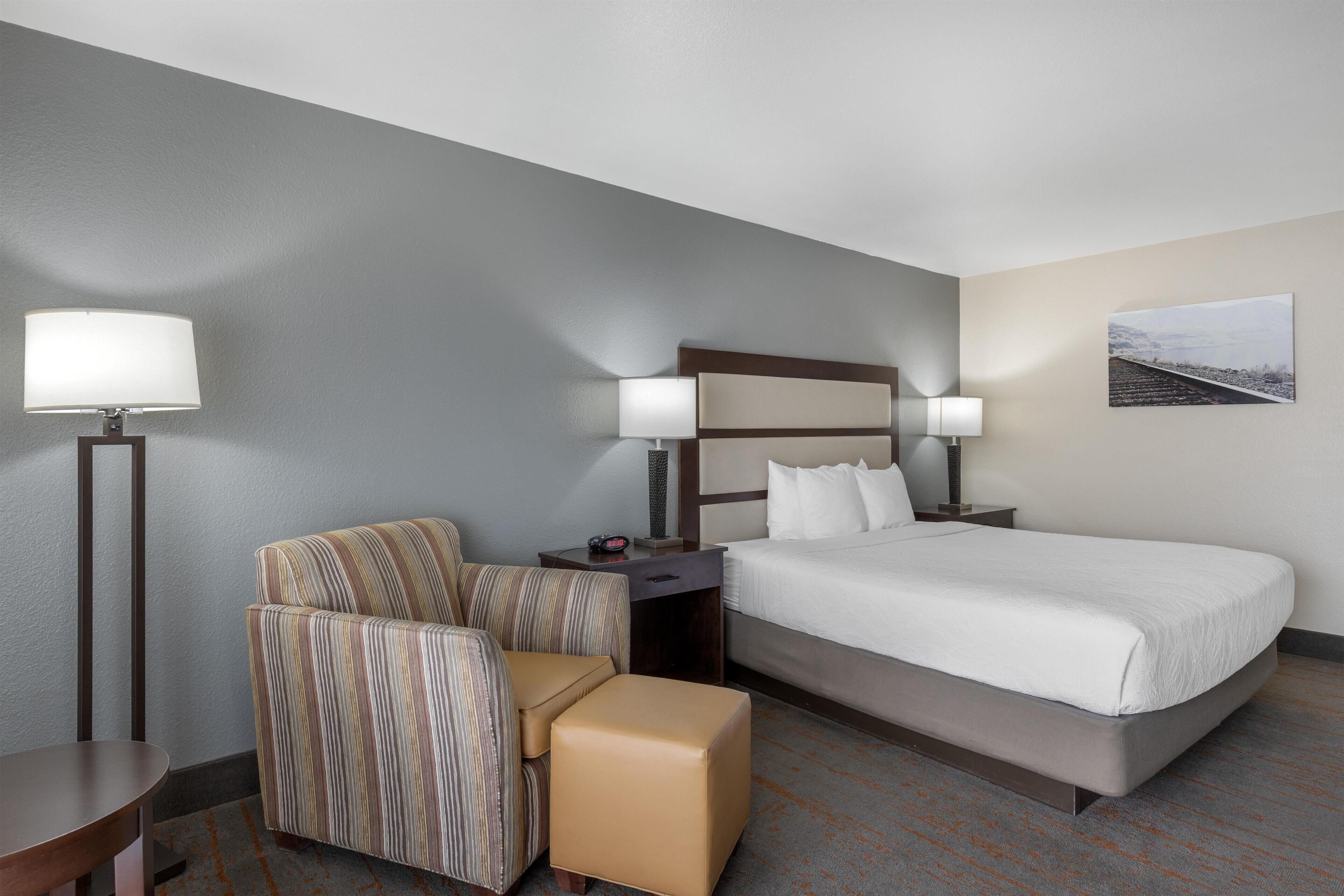 Una habitación de hotel con cama, una silla, una mesita, una mesita de noche y una lámpara.