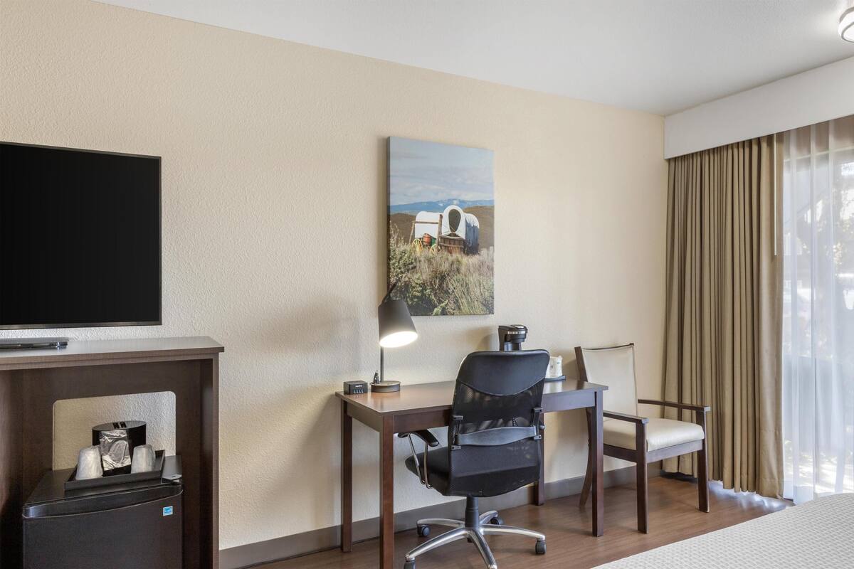 A hotel room with a desk, two chairs, a television, a lamp, and a painting on the wall.