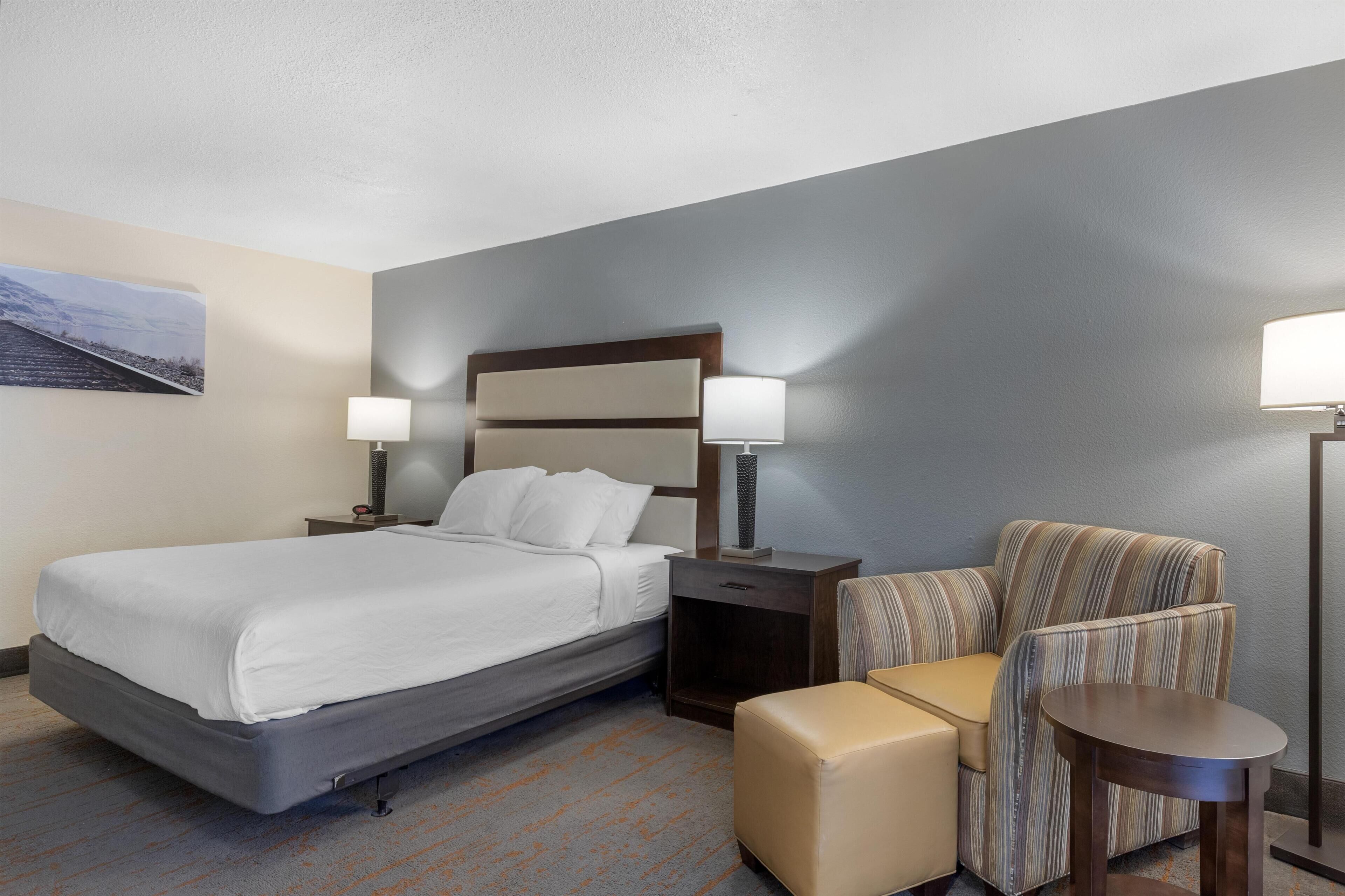 A hotel room with a bed, a nightstand with a lamp, a chair, a small table, and a wall-mounted picture.