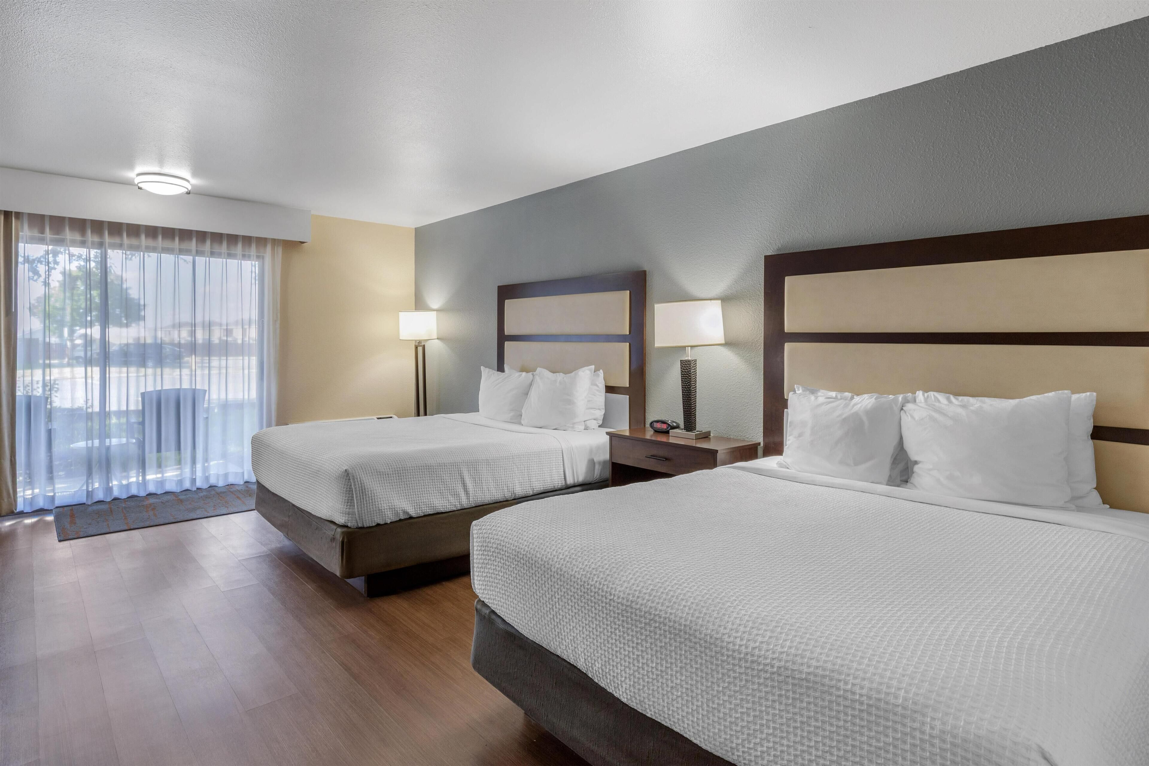 Habitación de hotel con dos camas, un ventanal con cortinas, una mesita de noche con lámpara y una lámpara del techo.
