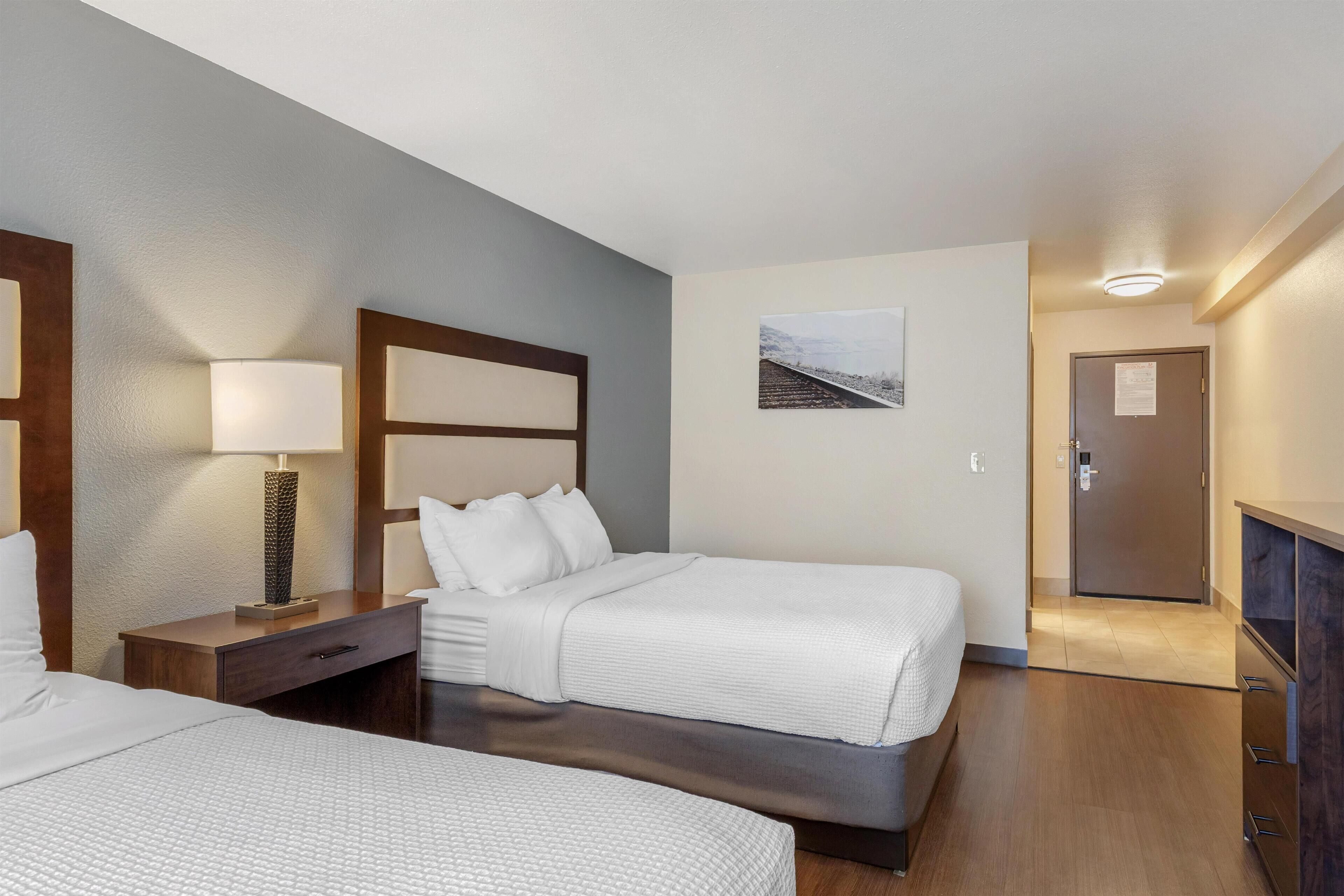 A hotel room with two beds, a nightstand, a lamp, a picture on the wall, and a door leading to a hallway.