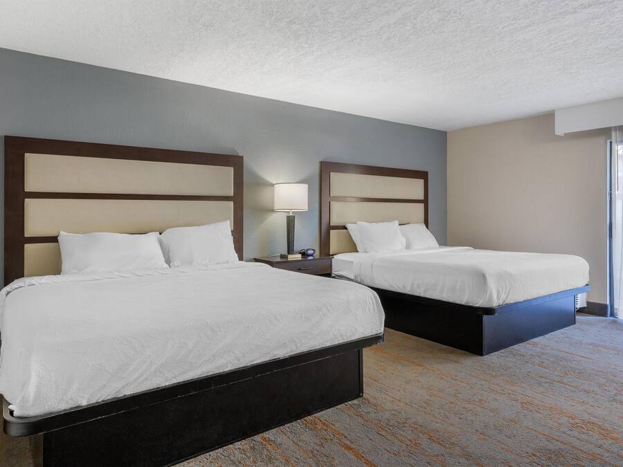 Habitación de hotel con dos camas, una mesita de noche, una lámpara y un ventanal con cortinas.