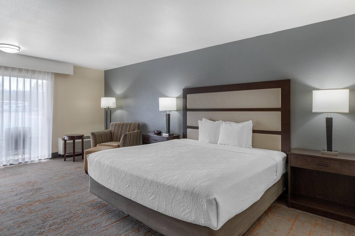 Une chambre d’hôtel avec un grand lit, deux tables de chevet avec des lampes, une chaise, une petite table et une grande fenêtre avec des rideaux.