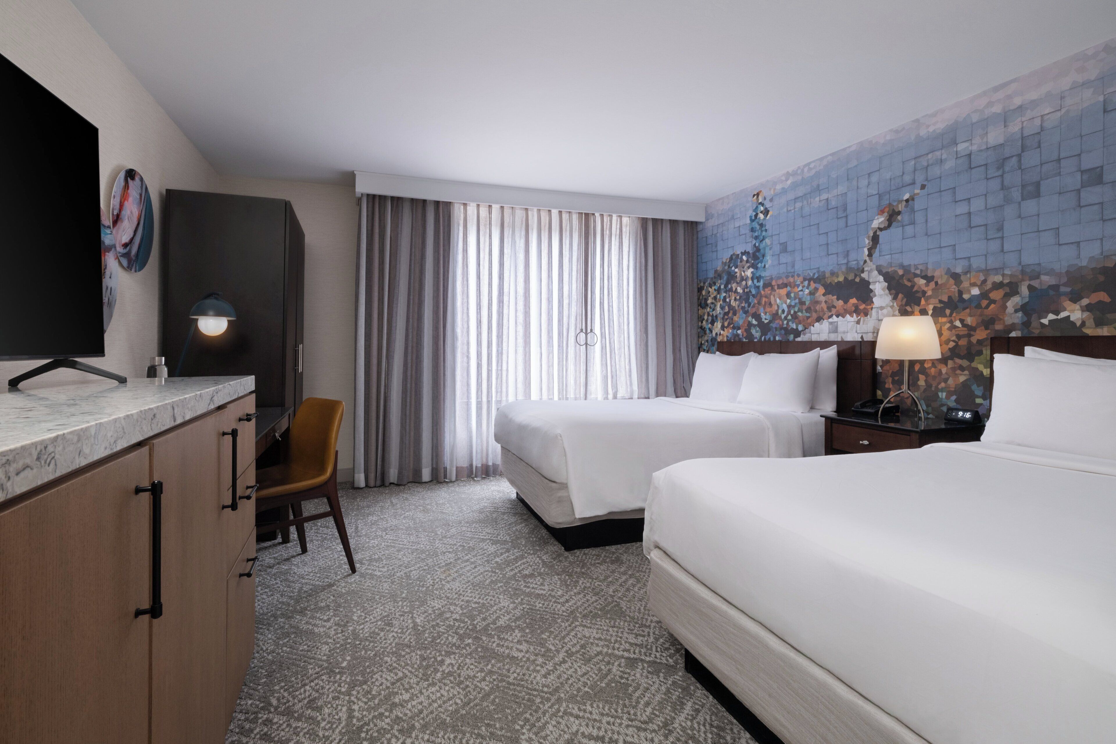 A hotel room with a bed, a desk with a chair, a desk lamp, a TV, and a large window with curtains.