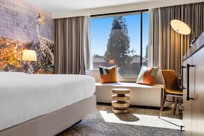 Quarto de hotel com uma cama grande, um sofá com almofadas coloridas, uma mesa de cabeceira de madeira, uma cadeira e uma janela ampla com vista para árvores e edifícios.