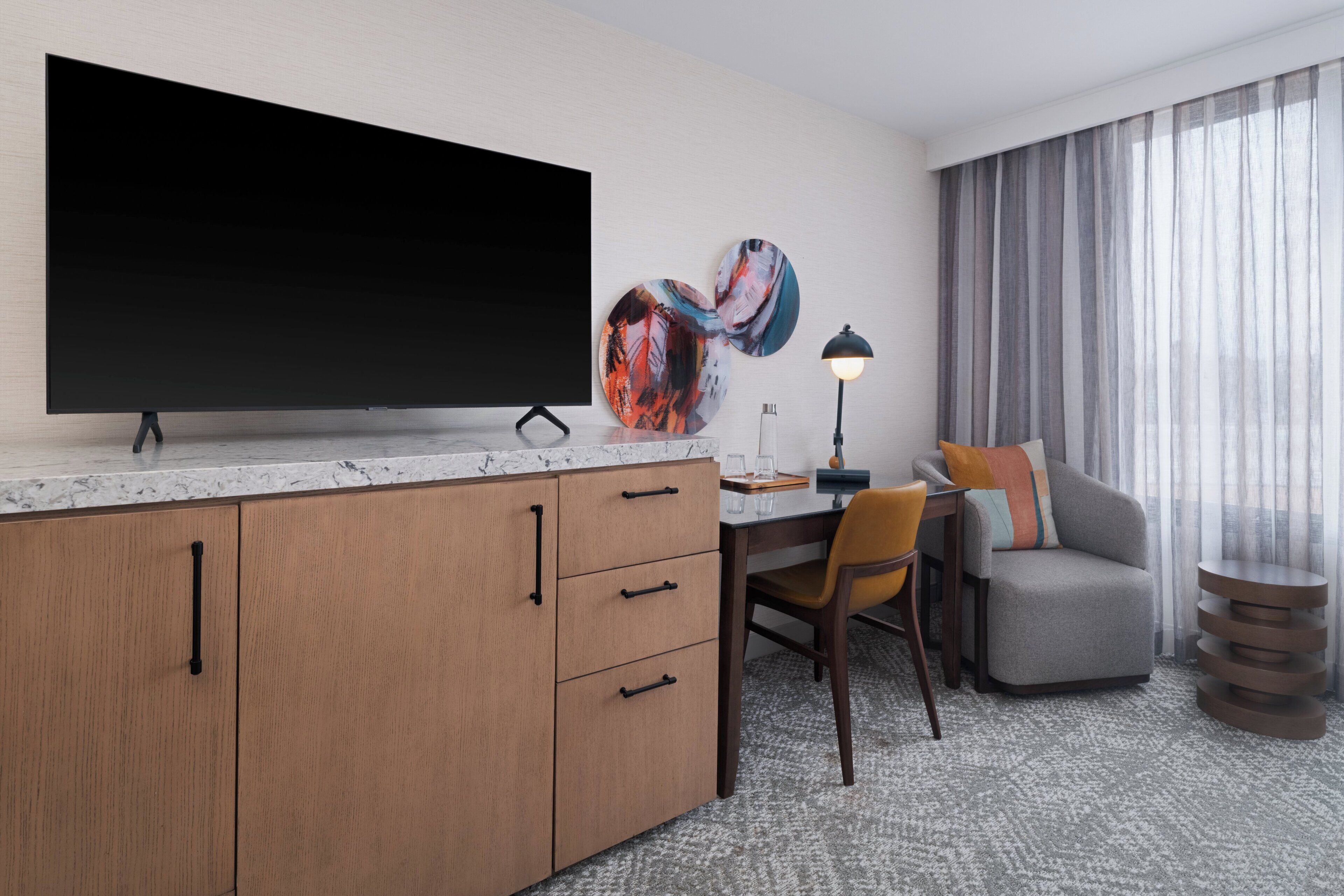 A modern hotel room with a large flat-screen TV, a desk with a chair, a sofa, and a decorative lamp.